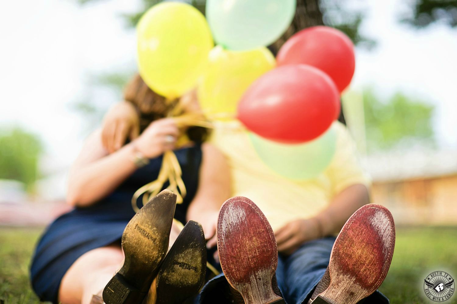 guy and gal sit next to each other with yellow, red and green balloons in front of their faces with shoes visible at the bluebonnet festival in burnett in this engagement image 